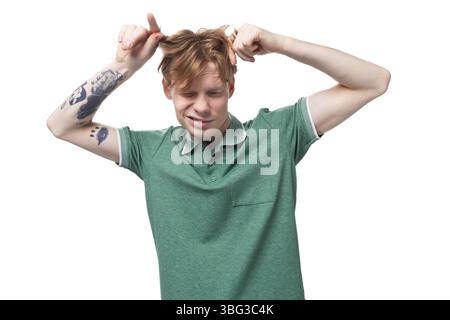 Il giovane ed elegante europeo con i capelli rossi e un tatuaggio sull'avambraccio indossa una t-shirt verde che gli tocca i capelli Foto Stock