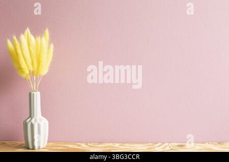 Vaso di prugnole gialle (Lagurus ovatus) fiori secchi su tavola di legno. Sfondo rosa Foto Stock