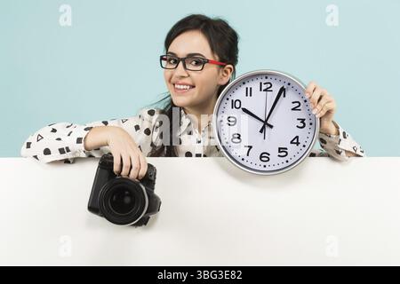 Ritratto di una giovane e attraente fotografa con camicia bianca e occhiali che tengono la macchina fotografica e osservano lo striscione bianco isolato su sfondo blu wi Foto Stock