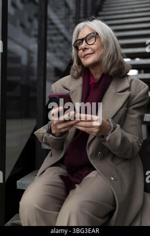 Una donna più anziana vestita con un cappotto sofisticato si siede comodamente su una scala, impegnata con il suo smartphone, mostrando un atteggiamento rilassato e contenuto Foto Stock