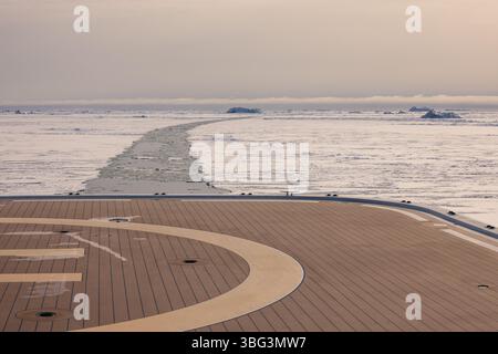 Vista dall'eliporto della nave che attraversa il ghiaccio marino vicino all'isola di Disko, in Groenlandia, con sentieri visibili e iceberg lontani. Foto Stock