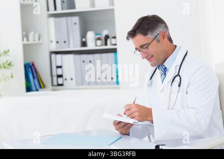 Maschio medico in bianco cappotto scrittura note su appunti a scrivania medica con stetoscopio, copia spazio Foto Stock