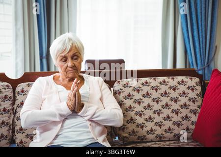 Donna anziana seduta su un divano floreale a casa con cuscino con accento rosso e tende blu Foto Stock