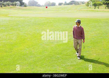 Golfista che cammina attraverso il vivace fairway verde verso la bandiera rossa, tenendo il putter con un guanto rosso Foto Stock