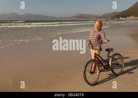 Donna anziana in piedi sul litorale sabbioso tenendo in mano una bicicletta rossa accanto alle onde dell'oceano, spazio fotocopie Foto Stock
