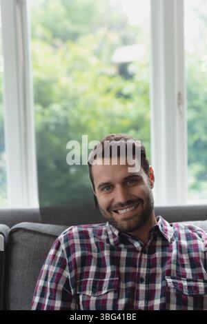 Uomo barbuto seduto su un divano grigio nel soggiorno sorridente davanti a una grande finestra con una camicia a quadri Foto Stock