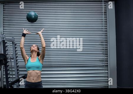 Donna ispanica che lancia una palla medica verde scuro sopra la testa in palestra con portapacchi e porta arrotolabile Foto Stock