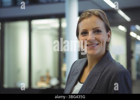 Donna che indossa un blazer e una blusa grigi in piedi vicino a una stanza con pareti in vetro in ufficio, spazio per le copie Foto Stock