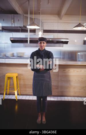 Chef indiano maschile in piedi al banco di legno sotto il cappuccio in acciaio inossidabile con cappotto nero e grembiule a righe Foto Stock