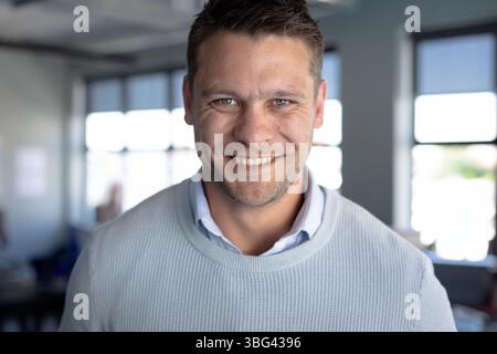 Uomo di 30 anni che indossa un maglione azzurro sorridente alla macchina fotografica in ufficio con scrivanie e grandi finestre Foto Stock