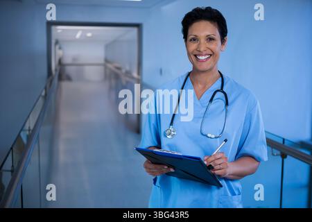 Infermiera donna che indossa scrub che tiene gli appunti e la penna nella sala dell'ospedale con lo stetoscopio, copia spazio Foto Stock