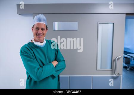 Uomo in scrub verdi, cappuccio chirurgico, maschera in piedi nella sala dell'ospedale dalla porta della barella, copia spazio Foto Stock