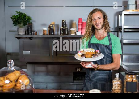Uomo sorridente, che tiene la torta sul piedistallo bianco con grembiule a righe dietro il bancone della panetteria Foto Stock