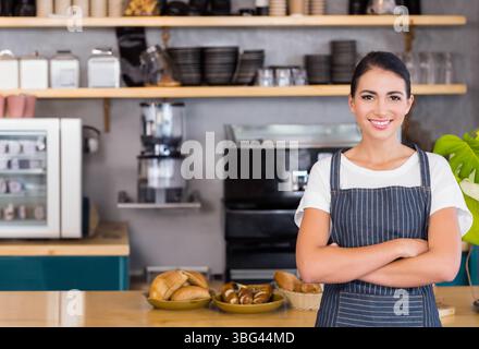 Barista donna in piedi dietro il bancone di legno che espone vassoi di pasticceria e panini al bar, spazio per le copie Foto Stock