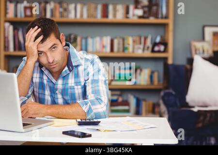 Uomo che lavora su documenti finanziari alla scrivania dell'ufficio con computer portatile, smartphone e documenti Foto Stock