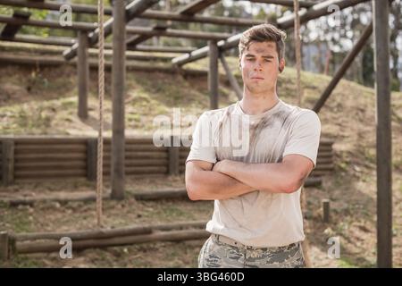 Apprendista militare maschile in piedi su un percorso ad ostacoli in legno con pantaloni camo, camicia fangosa e auricolari Foto Stock