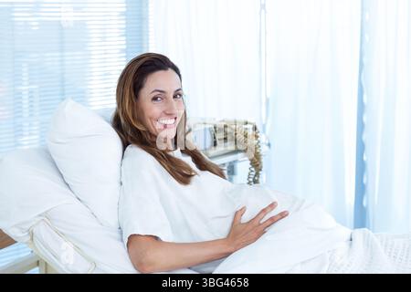 Donna incinta reclinata sul letto d'ospedale che indossa l'abito da ospedale, tenendo l'addome con il monitor nelle vicinanze Foto Stock