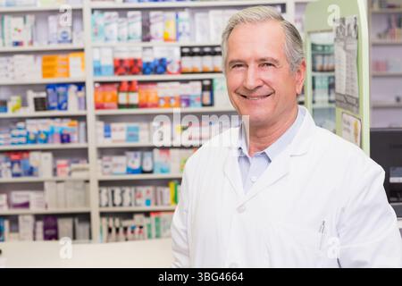 Farmacista maschile anziano in piedi dietro il banco della farmacia da scaffali della medicina, tabellone informativo, spazio di copia Foto Stock