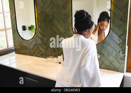 Donna afroamericana in accappatoio bianco che applica siero facciale al ripiano del bagno con specchi Foto Stock