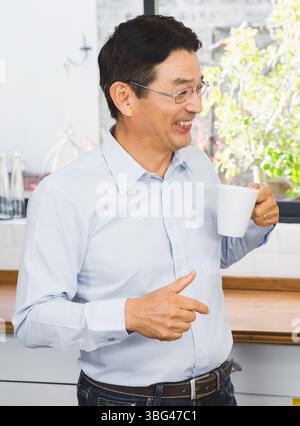 Un uomo giapponese anziano sorridente e tiene in mano una tazza da caffè bianca nella cucina casalinga sul piano di legno Foto Stock