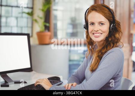 Lavoratrice in ufficio che indossa la cuffia con tastiera e mouse sulla scrivania vicino alla finestra con il monitor Foto Stock