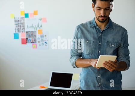 Un professionista del Medio Oriente che esamina un tablet in un ufficio moderno con collage di appunti e notebook Foto Stock