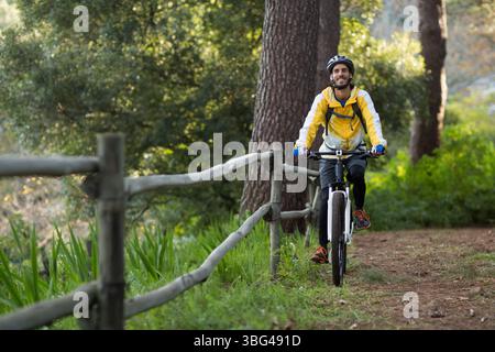 Uomo in bicicletta in mountain bike con giacca gialla e casco lungo il sentiero boscoso, oltre la recinzione di legno Foto Stock