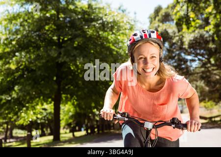 Ciclista donna di mezza età che pedala lungo il percorso del parco indossando un casco rosso e bianco, spazio fotocopie Foto Stock