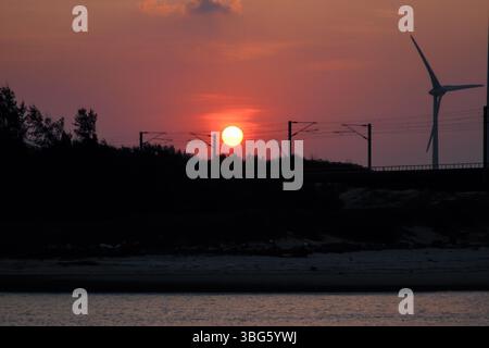Uno splendido tramonto dipinge il cielo in sfumature di rosso e arancione sopra Houlong Port, Miaoli County, Taiwan. Una turbina eolica e le linee aeree ferroviarie sono S Foto Stock