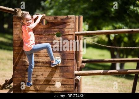 Bambina che si arrampica sulla parete del parco giochi in legno utilizzando le maniglie circolari e il ponte di corda nel parco, spazio fotocopie Foto Stock
