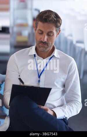 Uomo di mezza età che scrive sugli appunti seduto nella sala riunioni con sedie e badge con cordone Foto Stock
