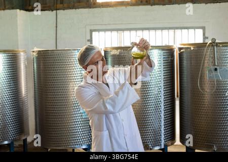 Uomo di mezza età che indossa un camice da laboratorio che esamina il liquido verde nel matraccio in un impianto di lavorazione con serbatoi Foto Stock