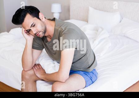 Un uomo adulto seduto sul bordo di un letto non realizzato nella camera da letto illuminata, tenendo la testa accanto alla lampada Foto Stock