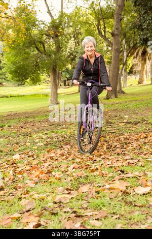 Donna anziana in bicicletta in autunno attraverso il parco su una bicicletta viola che indossa una giacca nera con auricolari Foto Stock