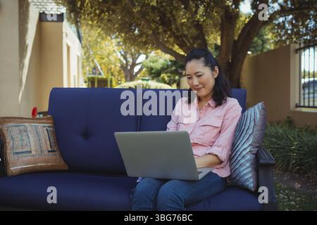 Donna coreana adulta matura seduta su un divano blu con laptop grigio argento con cuscini e spazio per fotocopie Foto Stock