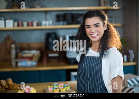Donna afro-americana sorridente che serve cupcake dietro il bancone in una caffetteria con grembiule e spazio per le copie Foto Stock