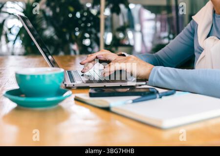 Le mani di una donna anziana che scrive su un computer portatile in un accogliente bar con notebook e caffè Foto Stock