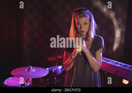 Cantante femminile che si esibisce su un piccolo palco sotto riflettori colorati con microfono e kit batteria Foto Stock