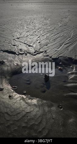 La marea si ritira, lasciando una piccola e incontaminata piscina d'acqua sulla spiaggia di sabbia, che riflette il cielo. Al centro esatto si distingue una pietra liscia Foto Stock