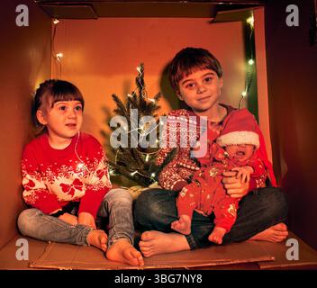 Tre bambini siedono in un'accogliente scatola di cartone decorata con luci fatate e un piccolo albero. Indossati con maglioni natalizi, creano un Natale caldo e fai da te Foto Stock