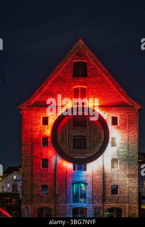 Un edificio in mattoni e' illuminato in modo drammatico con luci rosse, bianche e blu, caratterizzato da un grande anello nero sospeso al centro Foto Stock