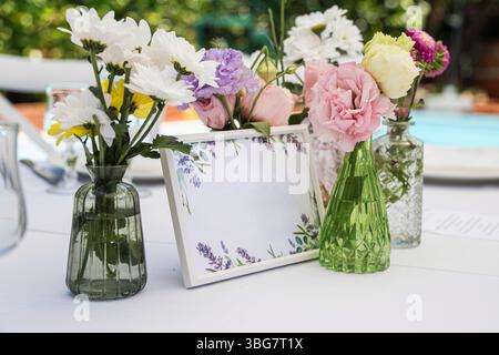 Elegante disposizione del tavolo con una cornice bianca in mockup circondata da piccoli vasi di colorati fiori primaverili. Perfetto per inviti di nozze, eventi ann Foto Stock