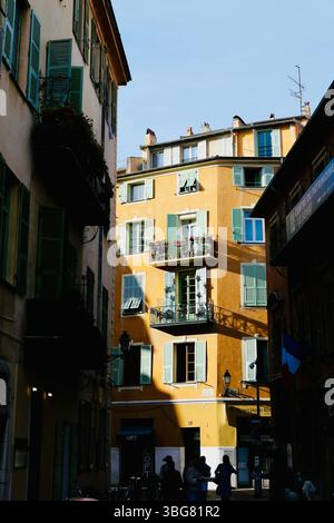 Edificio di appartamenti giallo illuminato dal sole nella città vecchia di Nizza, in Francia, visto da un vicolo ombreggiato con classiche persiane francesi e balconi. Foto Stock