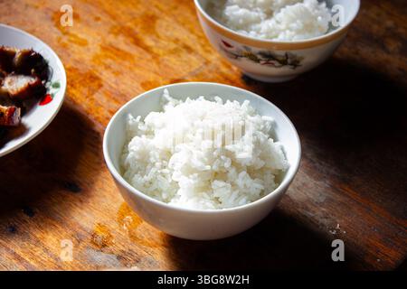 Primo piano di una ciotola di soffice riso bianco al vapore su un rustico tavolo di legno, con un'altra ciotola e un piatto di carne sfocato sullo sfondo. Foto Stock