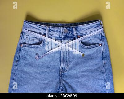 Jeans blu femminili con metro a nastro piatto su sfondo giallo. Concetto di perdita di peso, sovrappeso e stile di vita sano. Foto Stock
