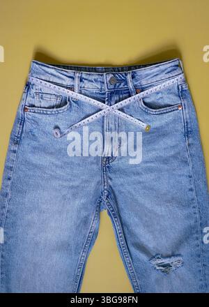 Jeans blu femminili con metro a nastro piatto su sfondo giallo. Concetto di perdita di peso, sovrappeso e stile di vita sano. Foto Stock