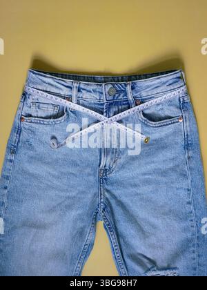 Jeans blu femminili con metro a nastro piatto su sfondo giallo. Concetto di perdita di peso, sovrappeso e stile di vita sano. Foto Stock