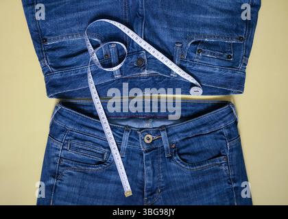 Due jeans blu femminili di dimensioni diverse con metro a nastro piatto su sfondo giallo. Concetto di perdita di peso, sovrappeso e stile di vita sano. Foto Stock