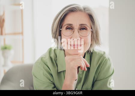 Affascinante donna matura con capelli grigi che indossa gli occhiali da vista seduti comodamente in casa e che proietta sicurezza e felicità Foto Stock