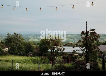 Vista sulla campagna pastorale con verdi colline, alberi e tetti sotto cieli nuvolosi. Villaggio serbo nella stagione primaverile. Foto Stock